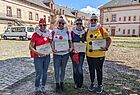 Vier Personen stehen nebeneinander in einem Innenhof mit historischen Gebäuden und tragen bunte Clownskostüme mit großen weißen Rüschenkragen, Pompons und passenden Mützen. Zwei sind in Rot-Weiß, einer in Blau-Gelb und einer in Gelb-Blau gekleidet. Alle tragen Sonnenbrillen und halten Mappen mit der Aufschrift „Die Mainzer Hofsänger“. Die Stimmung ist fröhlich und die Sonne scheint.