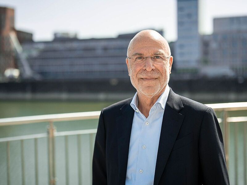 Älterer Mann mit Brille steht lächelnd im Freien vor einem Geländer; im Hintergrund befinden sich moderne Gebäude an einem Gewässer. Er trägt ein dunkles Sakko und ein helles Hemd.