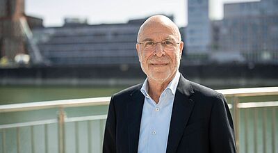 Älterer Mann mit Brille steht lächelnd im Freien vor einem Geländer; im Hintergrund befinden sich moderne Gebäude an einem Gewässer. Er trägt ein dunkles Sakko und ein helles Hemd.