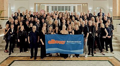 In Tunesien trafen sich die Franchise- Partner der Alltours Reisecenter zu ihrer Jahrestagung. Foto: Alltours Große Gruppenaufnahme von rund 80 Personen eines Alltours-Reisecenter-Teams, die in einer eleganten Hotel-Lobby auf einer Treppe stehen. Vorne halten mehrere Teilnehmende ein blaues Banner mit dem Alltours-Reisecenter-Logo. Alle blicken lächelnd in die Kamera.