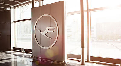 Eine Stele mit dem Logo von Lufthansa steht in einem Bürogebäude vor einer Glasfront, in der die Sonne hereinscheint