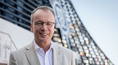 Ein Reederei-Manager Mitte 50 steht lächelnd an Deck eines Kreuzfahrtschiffes, im Hintergrund ist auf dem Schornstein unscharf das Logo von MSC Cruises zu erkennen. Der Mann hat kurzes braun, graues Haar, trägt eine Brille und ein hellbeiges Sakko auf weißem Oberhemd. 