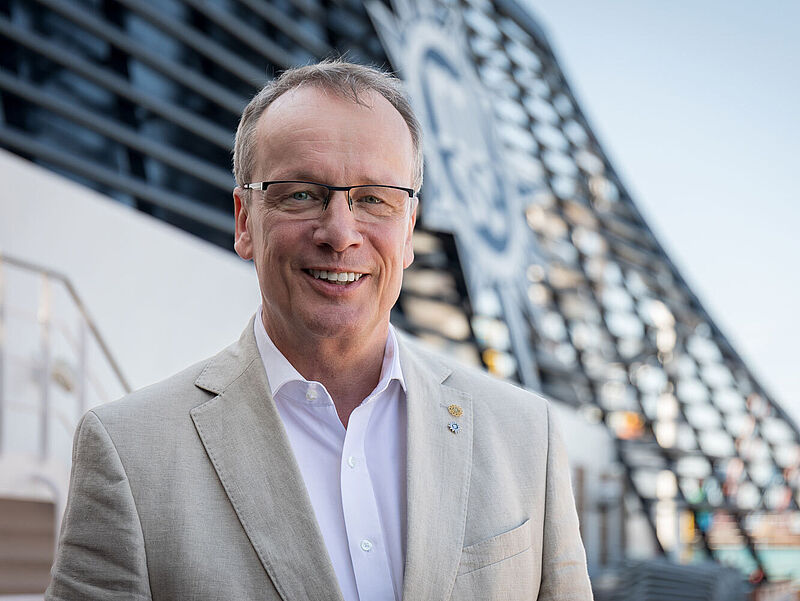 Ein Reederei-Manager Mitte 50 steht lächelnd an Deck eines Kreuzfahrtschiffes, im Hintergrund ist auf dem Schornstein unscharf das Logo von MSC Cruises zu erkennen. Der Mann hat kurzes braun, graues Haar, trägt eine Brille und ein hellbeiges Sakko auf weißem Oberhemd. 