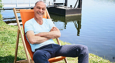 Der Schauspieler Martin Brambach, mit schütterem Haar und strahlendem Lächeln, sitzt in Freizeitkleidung in einem Liegestuhl an einem See und schaut in die Kamera. Die Sonne scheint.