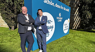 Wollen Holiday Land unter anderem noch digitaler aufstellen: Geschäftsführer Detlef Schroer und Michael Sawang Zwei Männer mittleren Alters in legerer Business-Kleidung stehen auf einem Rasenstück vor einem Aufsteller der Reisebüro-Kette Holiday Land an einem sonnigen Tag.