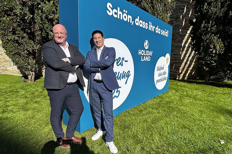 Wollen Holiday Land unter anderem noch digitaler aufstellen: Geschäftsführer Detlef Schroer und Michael Sawang Zwei Männer mittleren Alters in legerer Business-Kleidung stehen auf einem Rasenstück vor einem Aufsteller der Reisebüro-Kette Holiday Land an einem sonnigen Tag.