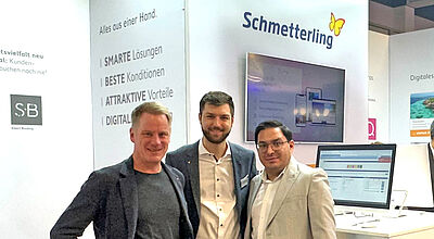 Drei Männer posieren lächelnd für ein Foto auf der Tourismusmesse ITB 2025 am Stand des Unternehmens Schmetterling. Im Hintergrund sind Monitore, Informationswände und weitere Messebesucher zu sehen. Der Messestand ist modern gestaltet mit dem Logo und Slogan des Unternehmens.