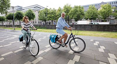 Auch zusätzliche Radtouren bietet Amadeus Flusskreuzfahrten im nächsten Jahr an. Foto: Lüftner Cruises Eine Frau und ein Mann mittleren Alters fahren mit City Bikes über eine asphaltierte Uferpromenade, im Hintergrund stehen grüne Bäume und graue Bürogebäude.