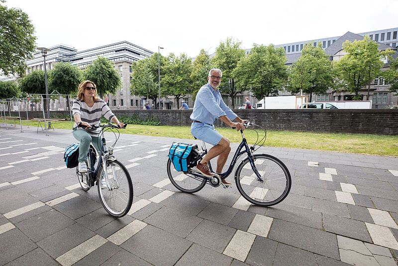 Auch zusätzliche Radtouren bietet Amadeus Flusskreuzfahrten im nächsten Jahr an. Foto: Lüftner Cruises Eine Frau und ein Mann mittleren Alters fahren mit City Bikes über eine asphaltierte Uferpromenade, im Hintergrund stehen grüne Bäume und graue Bürogebäude.