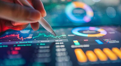 Durch einen automatisierten Datenaustausch werden bei Traffics künftig nicht verfügbare Hotels in Echtzeit aussortiert. Eine Hand hält einen digitalen Stift über einem leuchtenden Tablet, auf dem farbige Diagramme, Kurvenverläufe und Datenvisualisierungen angezeigt werden.