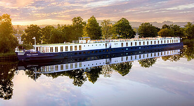 Nah an einem von Bäumen in sattem Grün gesäumten Flussufer liegt ein Flusskreuzfahrtschiff, dessen blauer Rumpf sich auf der glatten Wasseroberfläche spiegelt. Die Szenerie ist in stimmungsvolles orangefarbenes Sonnenlicht getaucht.