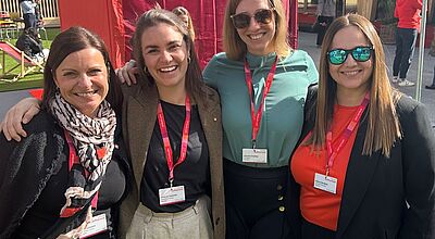 Vier lächelnde Frauen stehen nebeneinander und posieren für ein Foto im Freien. Sie tragen Namensschilder an roten Lanyards um den Hals. Im Hintergrund ist ein rotes Zelt mit der Aufschrift „Switzerland“ zu sehen.