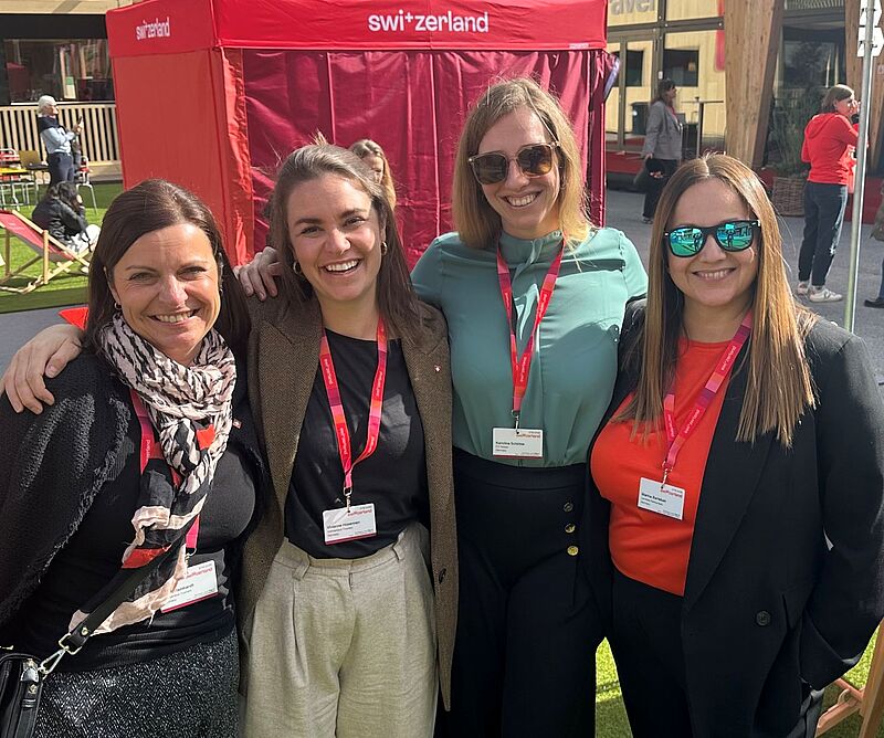 Vier lächelnde Frauen stehen nebeneinander und posieren für ein Foto im Freien. Sie tragen Namensschilder an roten Lanyards um den Hals. Im Hintergrund ist ein rotes Zelt mit der Aufschrift „Switzerland“ zu sehen.