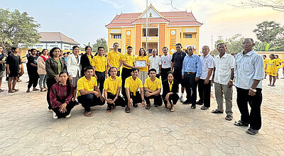 Dialogforum in Siem Reap, Kambodscha, mit lokalen Partnern des HVTO Sophy Village Free Education Program. Foto: Studiosus Eine große Gruppe von Menschen, bestehend aus Männern und Frauen unterschiedlichen Alters, steht auf einem Schulhof vor einem gelben Schulgebäude mit rotem Dach. Einige Personen tragen gelbe Shirts, andere sind formell gekleidet. In der Mitte der Gruppe wird eine Urkunde präsentiert. Im Hintergrund sind weitere Personen sowie Kinder in gelber Schuluniform zu sehen. Der Himmel ist leicht bewölkt und es herrscht eine freundliche Atmosphäre.