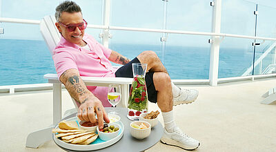 Mann in rosa Hemd entspannt auf Kreuzfahrtschiff-Balkon, genießt Snacks und Getränke mit Meerblick.