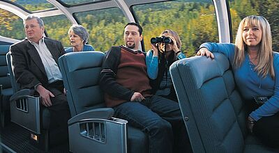 Die Zugreise „Goldener Ahorn“ gehört zu den Klassikern bei Lernidee. Sie beinhaltet eine Fahrt im Zug The Canadian. Foto: Via Rail Canada Eine Gruppe von Menschen sitzt entspannt in einem Panoramawagen eines Zuges mit großen, gewölbten Glasfenstern, durch die man eine malerische Landschaft mit Wäldern und Bergen sehen kann. Eine Frau benutzt ein Fernglas, um die Aussicht zu betrachten. Die Atmosphäre wirkt ruhig und genussvoll, während der Zug durch die Natur fährt.