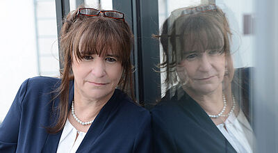 Frau im Business-Outfit mit Perlenkette und Brille, spiegelt sich nachdenklich in einer Glasfassade.