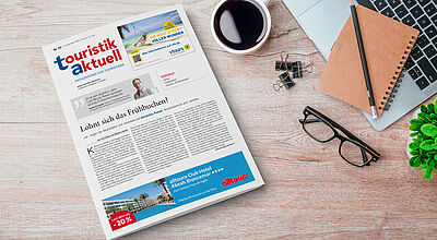 Auf einem Schreibtisch liegen eine Ausgabe der Fachzeitung touristik aktuell, eine Brille, ein Notizbuch mit Bleistift, eine Tasse Kaffee, Büroklammern, ein Laptop und eine kleine grüne Pflanze