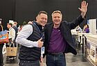 Zwei Männer zwischen Ständen lachen in die Kamera, der eine winkt