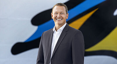 Ein Reederei-Manager Mitte 40 mit kurzen braunen Haaren und blauen Augen steht vor einem Kreuzfahrtschiff und blickt freudig lächelnd in die Kamera.