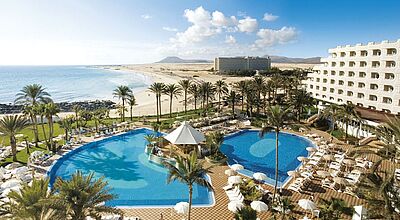 Ein weißes Hotelgebäude mit einem großen blauen Pool an einem Sandstrand