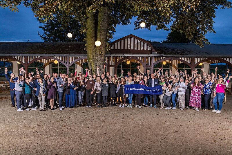 Eine große Gruppe Menschen mit einem Banner, auf dem Lufthansa City Center, steht lächelt in die Kamera. Die Veranstaltung findet im freien statt. Die Stimmung ist ausgelassen.