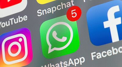 Nahaufnahme eines Smartphone-Bildschirms mit mehreren Social-Media-Apps. Im Fokus steht das grüne WhatsApp-Symbol mit einer roten Benachrichtigung („5“) oben rechts. Daneben sind die Logos von Instagram, YouTube, Snapchat und Facebook zu sehen.