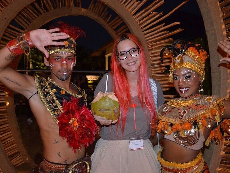 Teilnehmerin einer Dertour-Gruppenreise in der Dominikanischen Republik posiert bei einer Abendveranstaltung mit zwei traditionell gekleideten Tänzern in farbenfrohen Kostümen und hält eine Kokosnuss mit Logo – kulturelle Begegnung und Tourismus-Erlebnis in der Karibik.