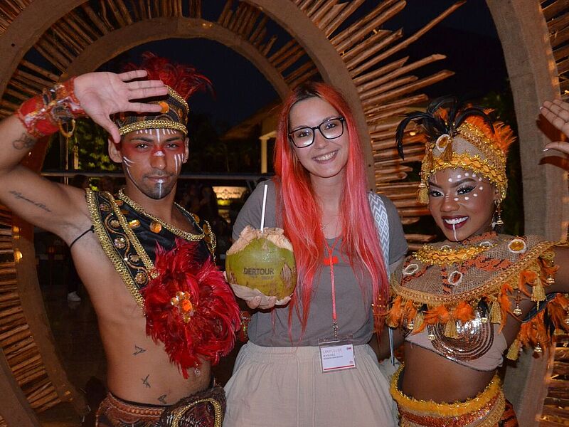 Teilnehmerin einer Dertour-Gruppenreise in der Dominikanischen Republik posiert bei einer Abendveranstaltung mit zwei traditionell gekleideten Tänzern in farbenfrohen Kostümen und hält eine Kokosnuss mit Logo – kulturelle Begegnung und Tourismus-Erlebnis in der Karibik.