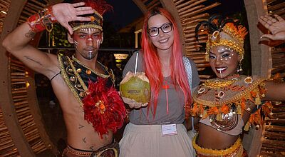 Teilnehmerin einer Dertour-Gruppenreise in der Dominikanischen Republik posiert bei einer Abendveranstaltung mit zwei traditionell gekleideten Tänzern in farbenfrohen Kostümen und hält eine Kokosnuss mit Logo – kulturelle Begegnung und Tourismus-Erlebnis in der Karibik.