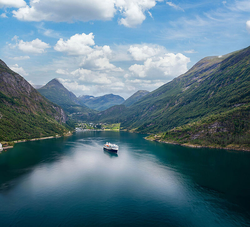 Ein Küstenkreuzfahrtschiff mit dunkelblaem Rumpf und weißem Aufbau befährt den berühmten Geirangerfjord in Norwegen. Am Ende des Fjord ist der gleichnamige Ort zu erkennen, links und rechts ragen bewaldete Berge empor. 
