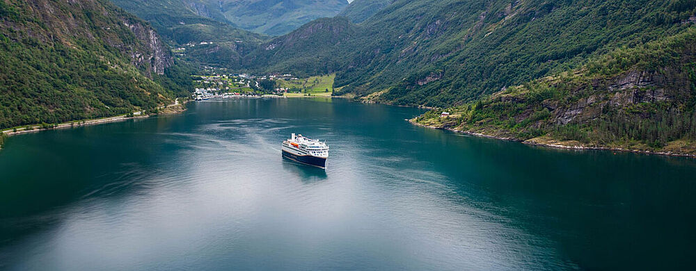 Ein Küstenkreuzfahrtschiff mit dunkelblaem Rumpf und weißem Aufbau befährt den berühmten Geirangerfjord in Norwegen. Am Ende des Fjord ist der gleichnamige Ort zu erkennen, links und rechts ragen bewaldete Berge empor. 
