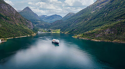 Ein Küstenkreuzfahrtschiff mit dunkelblaem Rumpf und weißem Aufbau befährt den berühmten Geirangerfjord in Norwegen. Am Ende des Fjord ist der gleichnamige Ort zu erkennen, links und rechts ragen bewaldete Berge empor. 