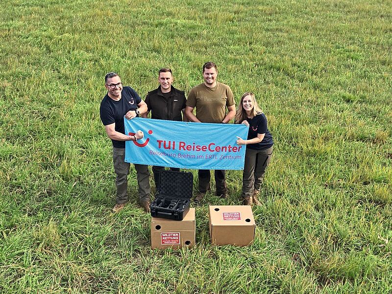 Matthias Brehm, Wolfgang Schmitt, Stephan Schmitt und ­Nicole Brehm (von links) beim Einsatz und auf der Suche nach Rehkitzenn auf einer Wiese mit einem Banner des Reisebüros in der Hand