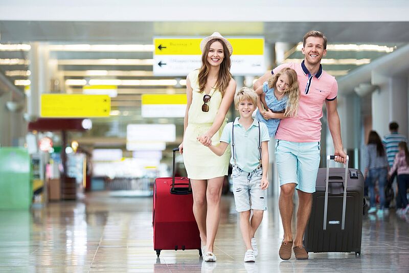 Eine fröhliche Familie mit zwei Kindern geht durch einen Flughafen. Die Mutter trägt ein hellgelbes Sommerkleid und zieht einen roten Rollkoffer. Der Vater trägt ein rosa Polohemd und zieht einen schwarzen Rollkoffer, während er die Tochter auf dem Arm trägt. Der Sohn läuft zwischen den Eltern und hält ihre Hände. Im Hintergrund sind Schilder für Wegweiser und öffentliche Verkehrsmittel zu sehen.
