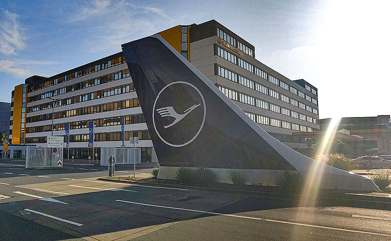 Lufthansa-Produkte über NDC zu buchen und zu modifizieren, soll jetzt deutlich einfacher werden Blick auf die Lufthansa-Basis am Flughafen Frankfurt mit einem Seitenleitwerk mit Lufthansa-Logo im Vordergrund im Gegenlicht der Sonne.