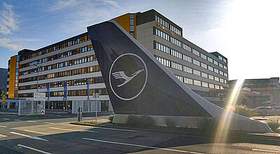 Lufthansa-Produkte über NDC zu buchen und zu modifizieren, soll jetzt deutlich einfacher werden Blick auf die Lufthansa-Basis am Flughafen Frankfurt mit einem Seitenleitwerk mit Lufthansa-Logo im Vordergrund im Gegenlicht der Sonne.