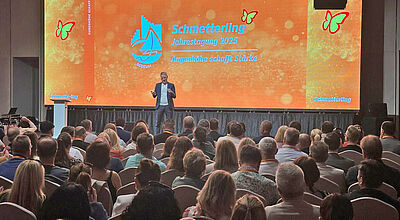 Konferenzraum mit Redner auf Bühne vor großer Leinwand. Logo "Schmetterling Jahrestagung 2023" und Motto "Augenhöhe schafft Stärke" sichtbar.