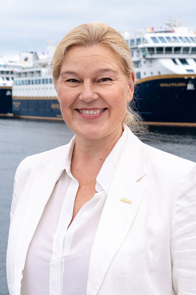 Eine Frau mittleren Alters mit blondem Pferdeschwanz und in weißer Bluse und weißem Sakko steht lächelnd an einem Kreuzfahrtpier. Hinter ihr ist ein Kreuzfahrtschiff zu erkennen.