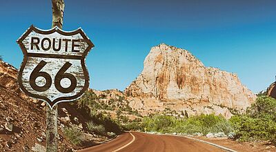 Bild von einer Straße unter blauem Himmel mit rötlichen Felsen im Hintergrund und einem Straßenschild links im Vordergrund, auf dem Route 66 geschrieben steht.