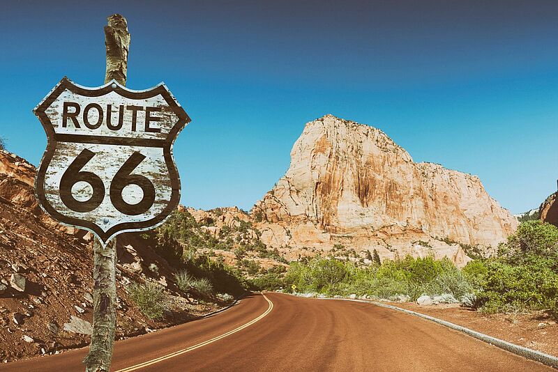 Bild von einer Straße unter blauem Himmel mit rötlichen Felsen im Hintergrund und einem Straßenschild links im Vordergrund, auf dem Route 66 geschrieben steht.