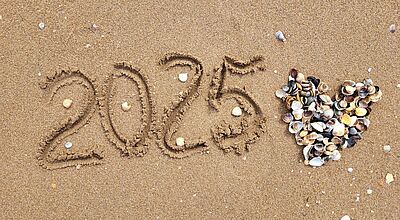 Am Strand ist die Zahl 2025 in den Sand geschrieben, rechts daneben ein Herz aus unterschiedlichen Muscheln gelegt