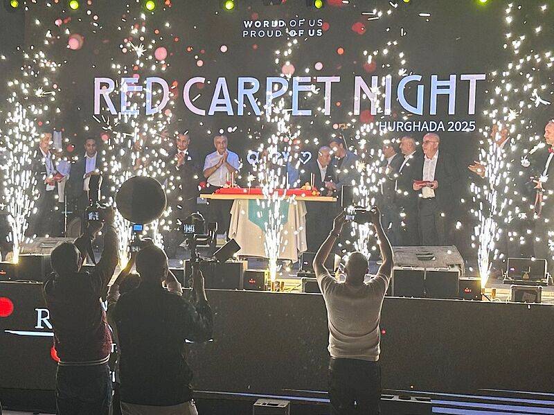 Der Galaabend zur Red Carpet Night fand im Rixos Magawish in Hurghada statt Bühnenaufnahme einer feierlichen Veranstaltung mit großem Feuerwerkseffekt. Auf der beleuchteten Bühne stehen mehrere Personen in formeller Kleidung hinter einem Tisch, während Funkenfontänen links und rechts hochschießen. Im Vordergrund fotografieren und filmen mehrere Personen die Szene. Auf der Bühnenrückwand steht groß „Red Carpet Night – Hurghada 2025“.