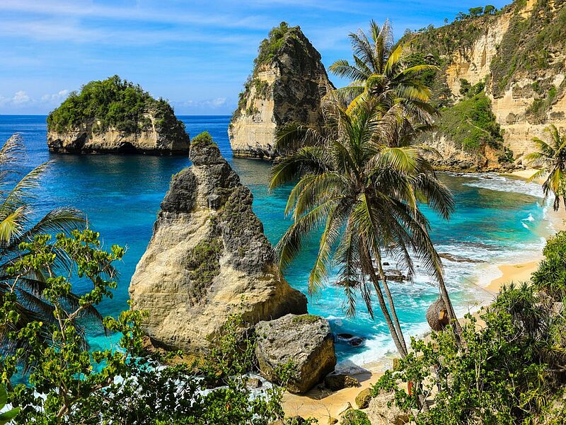Bali ist neben Sri Lanka neu im Asien-Portfolio von Coral Travel. Foto: EoNaYa/istock Tropische Küstenlandschaft mit türkisblauem Meer, markanten Felsformationen und steilen Klippen. Im Vordergrund stehen mehrere Palmen, dahinter liegen helle Sandstrände und große, von Vegetation bewachsene Felsen, die aus dem Wasser ragen.