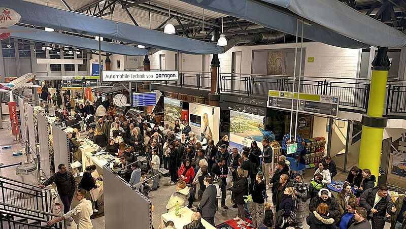 Die Aufnahme zeigt eine geschäftige Reisemesse in der Abflughalle des Flughafens Paderborn-Lippstadt. Zahlreiche Besucher strömen durch die Messegänge, während verschiedene Aussteller ihre Reiseangebote an Ständen präsentieren. Im Hintergrund sind Werbebanner von Reiseanbietern und Hotels zu sehen. Oberhalb der Szene hängen Wegweiser für Taxis, Busse und Hotels. Eine große Uhr sowie Anzeigetafeln mit Flugzeiten verstärken das Flughafenumfeld