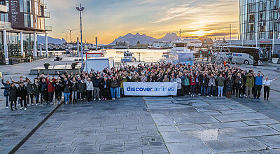 Rund 120 Reiseprofis, hier in Svolvaer, erleben gerade intensiv die Lofoten und andere norwegische Regionen. Foto: Dave Williams/Visit Lofoten/Discover Airlines Eine große Gruppe von über 100 Reisebüro-Mitarbeitern stehen auf dem zentralen Platz Stadtzentrum von Solvaer in der norwegischen Lofoten-Region vor einer Berg- und Hafenkulisse.