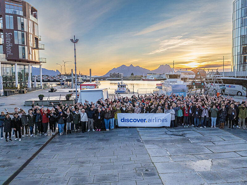 Eine große Gruppe von über 100 Reisebüro-Mitarbeitern stehen auf dem zentralen Platz Stadtzentrum von Solvaer in der norwegischen Lofoten-Region vor einer Berg- und Hafenkulisse.