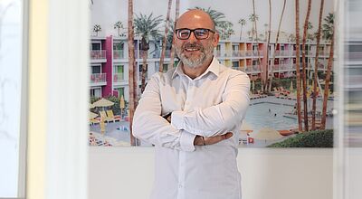 Ein Mann in weißem Hemd steht mit verschränkten Armen vor einem farbenfrohen Bild eines Hotels mit Palmen und Pool