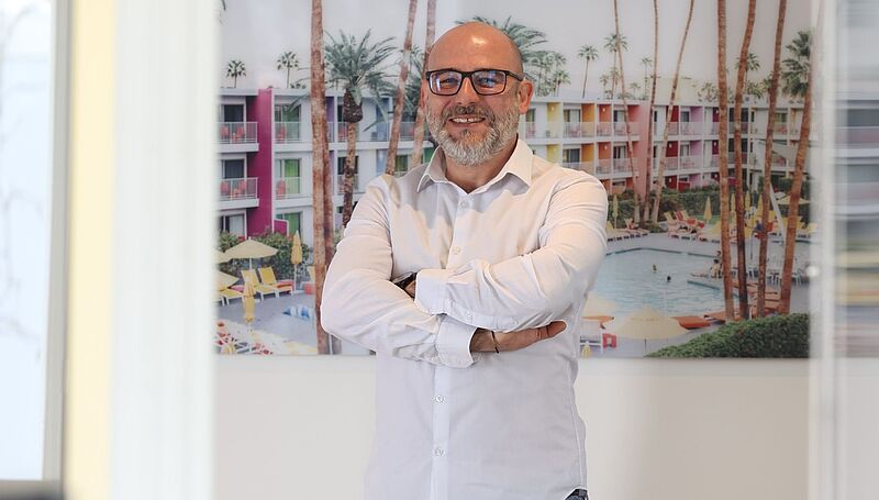 Ein Mann in weißem Hemd steht mit verschränkten Armen vor einem farbenfrohen Bild eines Hotels mit Palmen und Pool