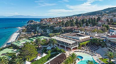 Luftaufnahme des Aldiana Rocca Nettuno Calabria: Das Resort liegt auf einem Felsplateau direkt an der kalabrischen Küste mit Blick auf das Tyrrhenische Meer. Zu sehen sind der weitläufige Poolbereich mit Palmen, das Hauptgebäude mit Restaurantterrasse sowie der flach abfallende Sandstrand und die Altstadt von Tropea im Hintergrund.